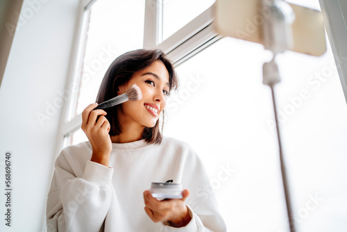 Young influencer applying blusher and filming through mobile phone at home