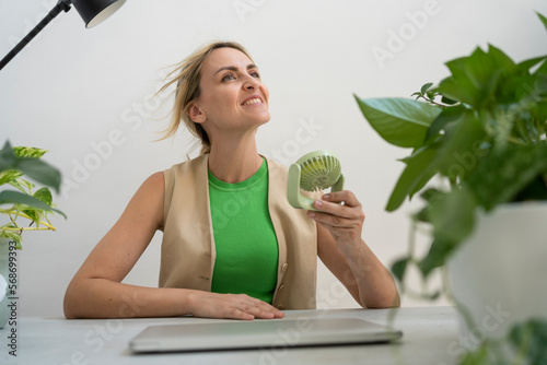 Happy freelancer with handheld motorized fan sitting at desk photo