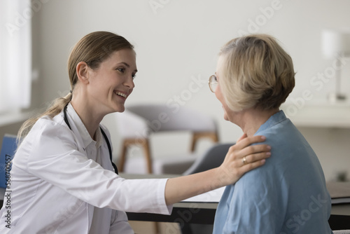 Positive physician woman talking to elderly patient, touching shoulder, smiling, giving support, comfort, medical advice. Senior lady visiting doctor for medical examination