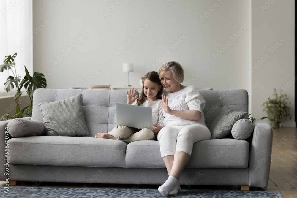 Cheerful granny and grandchild talking on video call, using laptop on couch together, waving hand at screen, enjoying domestic Internet communication, smiling, laughing Cheerful granny and grandchild talking on video call, using laptop on couch together, waving hand at screen, enjoying domestic Internet communication, smiling, laughing