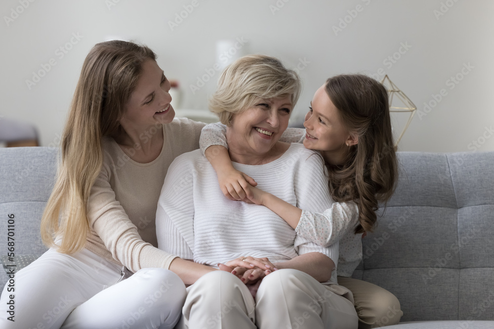 Sweet kid girl and mom visiting happy grandma, sitting on home sofa together, chatting, laughing, having fun, hugging with affection, enjoying family holiday, affection, relationship - obrazy, fototapety, plakaty 