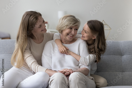 Sweet kid girl and mom visiting happy grandma, sitting on home sofa together, chatting, laughing, having fun, hugging with affection, enjoying family holiday, affection, relationship