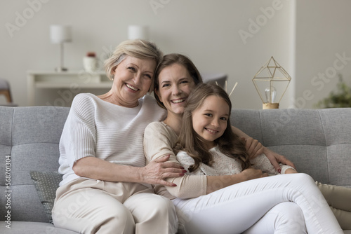 Happy joyful girls and women of three family generations relaxing on home sofa together, hugging, cuddling kid, looking at camera, laughing, smiling, enjoying leisure time, weekend