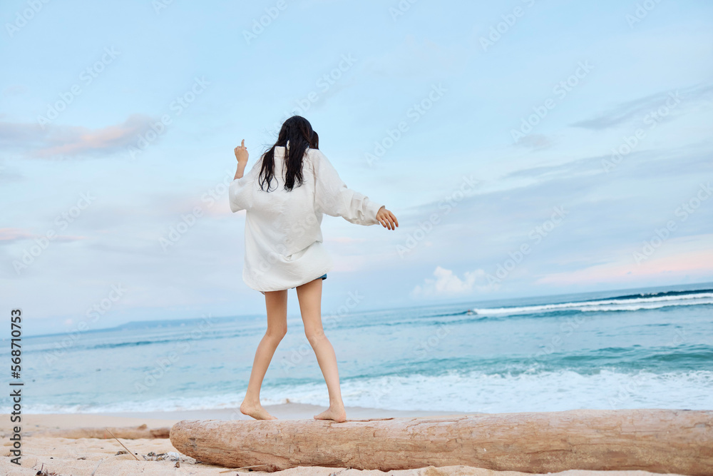 Happy tanned woman in white swimsuit shirt and denim shorts walks the beach on a log on the sand by the ocean with wet hair after swimming, sunset light in Bali Happy tanned woman in white swimsuit shirt and denim shorts walks the beach on a log on the sand by the ocean with wet hair after swimming, sunset light in Bali
