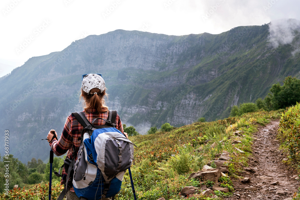 rear view of young woman in plaid shirt and cap with big backpack with trekking poles walking along mountain trail in summer hiking healthy active lifestyle looking at view copy space