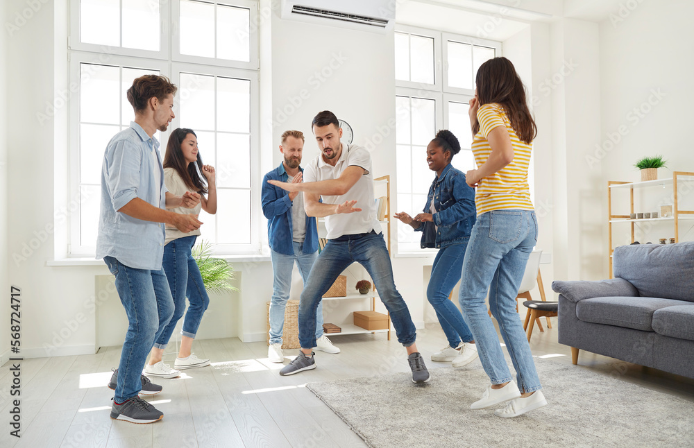 Young, cheerful and funny friends dancing and laughing while meeting at house party. Man dances funny in circle of his multiracial friends. Concept of friendship, home entertainment and people.