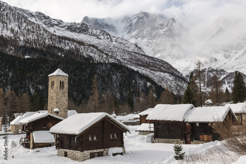 Snowy winter landscape of the Italian Alps with old fashioned wooden huts. Macugnaga, important ski resort in Piedmont with Monte Rosa partially enveloped by clouds. Anzasca valley, Italy