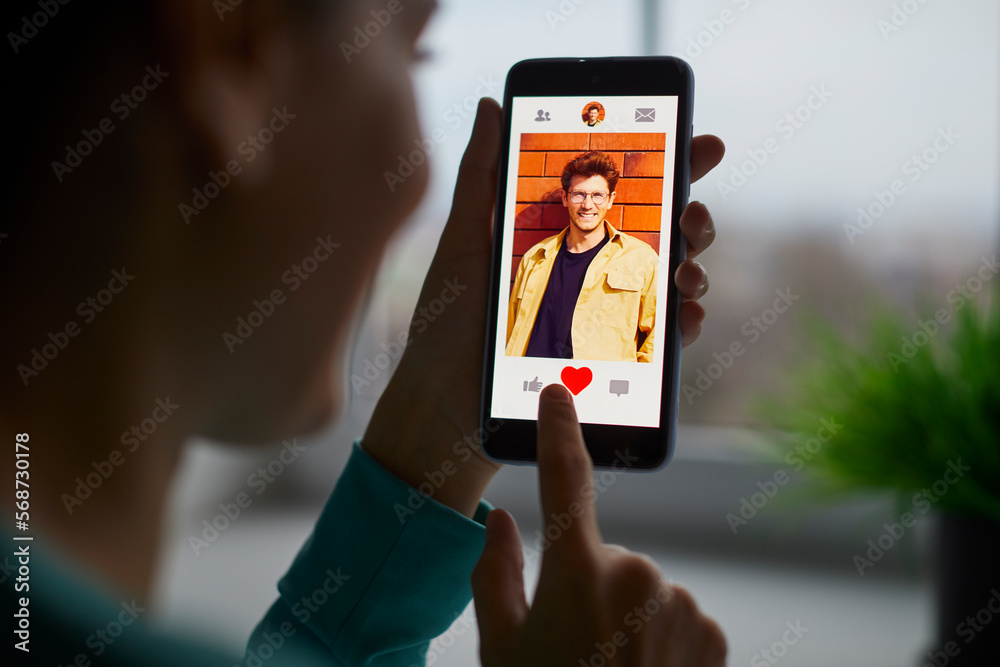 Portrait of a smiling young woman putting red heart button giving like to a cheerful handsome man on her mobile dating application. Find love and online dating concept. Selective focus.