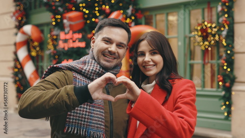 Portrait of the happy family couple making heart gesture with fingers in the decorated street, caucasian man and woman showing love hands, demonstrating sincere feelings together. Dating time