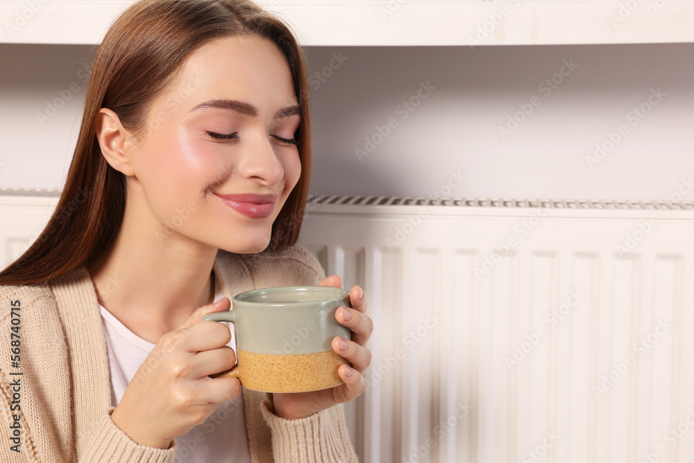 Woman holding cup with hot drink near heating radiator indoors