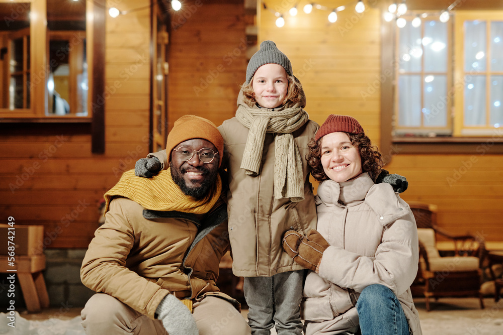 Cheerful intercultural mother and father in warm winterwear surrounding their happy cute little son while enjoying winter vacation