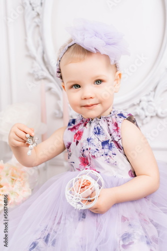 Baby girl elegant dress. A one-year-old girl in a puffy dress and a cute bow poses against the backdrop of a bright room with a dressing table and flowers.
