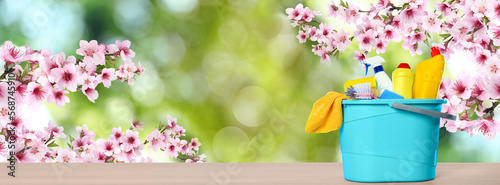Fototapeta Naklejka Na Ścianę i Meble -  Spring cleaning. Bucket with detergents and tools on wooden surface under blossoming tree against blurred green background, space for text. Banner design