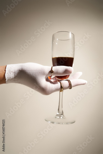 Cropped close-up shot of a woman's hand in a white glove with a glass of red wine. Emphasis is on elegant gloves made of elastic openwork fine knit fabric.  Side view.
