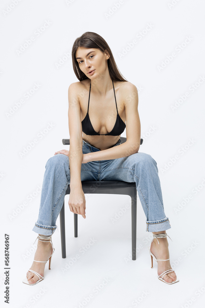 Stylish young woman in jeans black top and heels sitting on a chair in the studio on a white background Stylish young woman in jeans black top and heels sitting on a chair in the studio on a white background