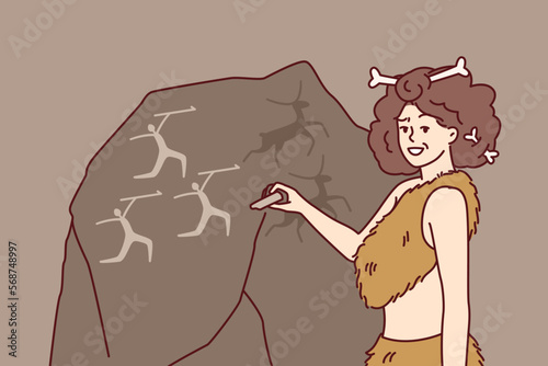 Ancient woman in clothing made from animal skins and bones in hair makes rock paintings. Girl depicts people hunting deer on giant stone and smiling looks at screen