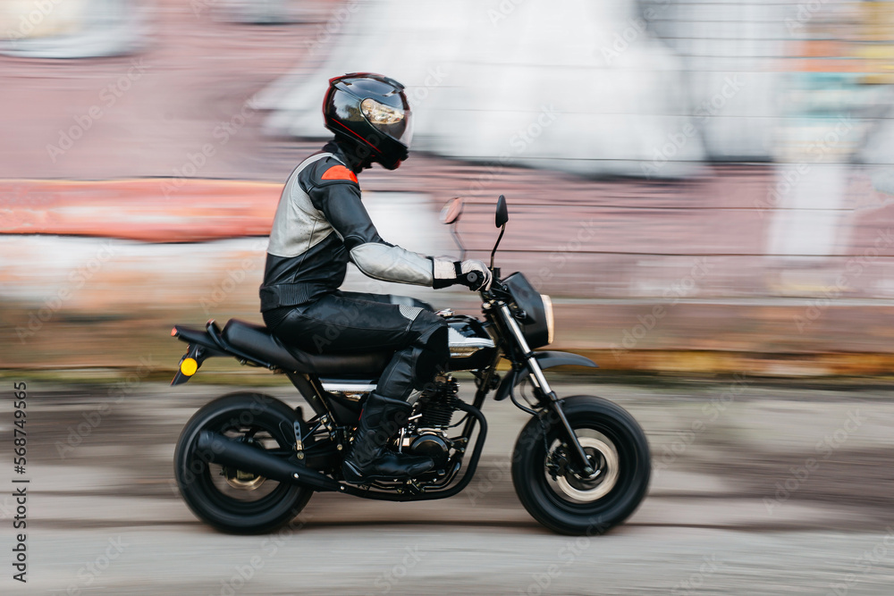 Motorcyclist in full black leathers and helmet riding fast on a background of the wall with graffiti