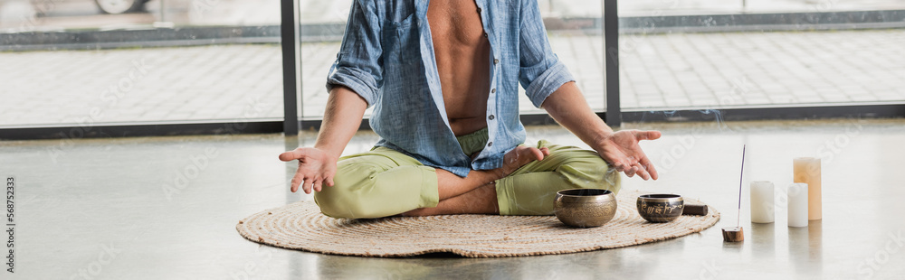 cropped view of man sitting in lotus pose while meditating near Tibetan singing bowls in yoga studio, banner.