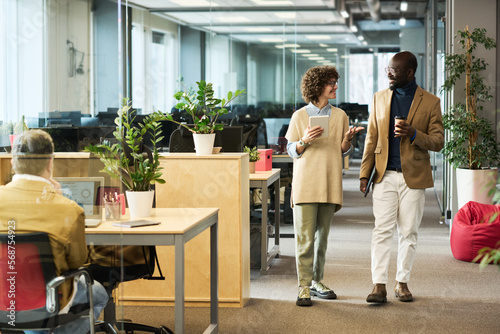 Woman with tablet and her colleague with cup of coffee discussing main working points while moving along rows of desks in openspace office