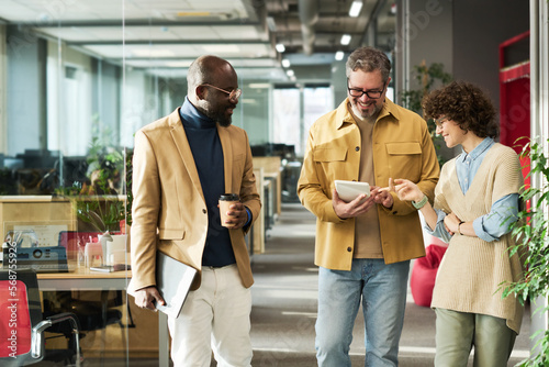 Group of three intercultural colleagues in casual attire discussing online data or video on screen of tablet held by mature male manager