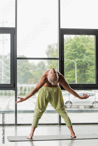 full length of shirtless man in green pants doing backside bend in yoga studio.