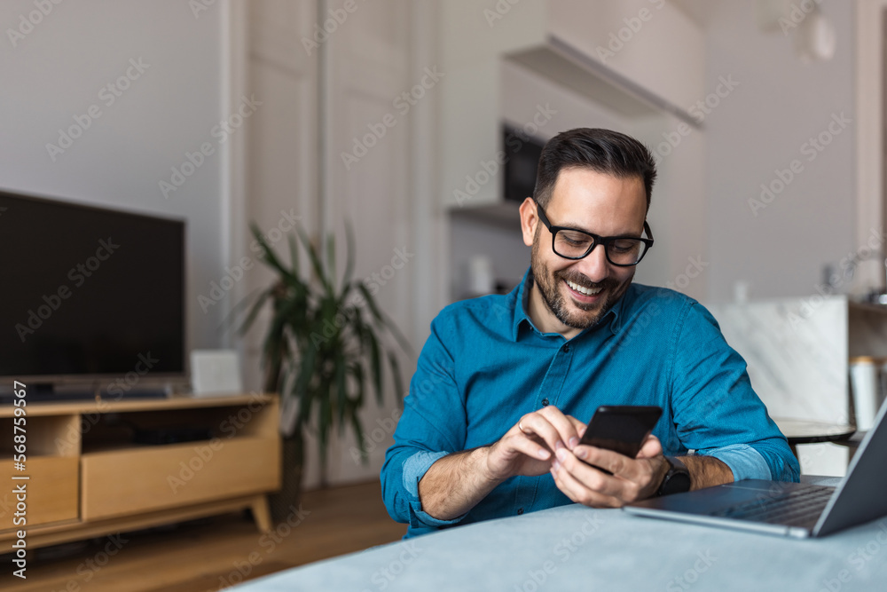 Smiling businessman using a mobile phone, sitting in front of a laptop, indoors.