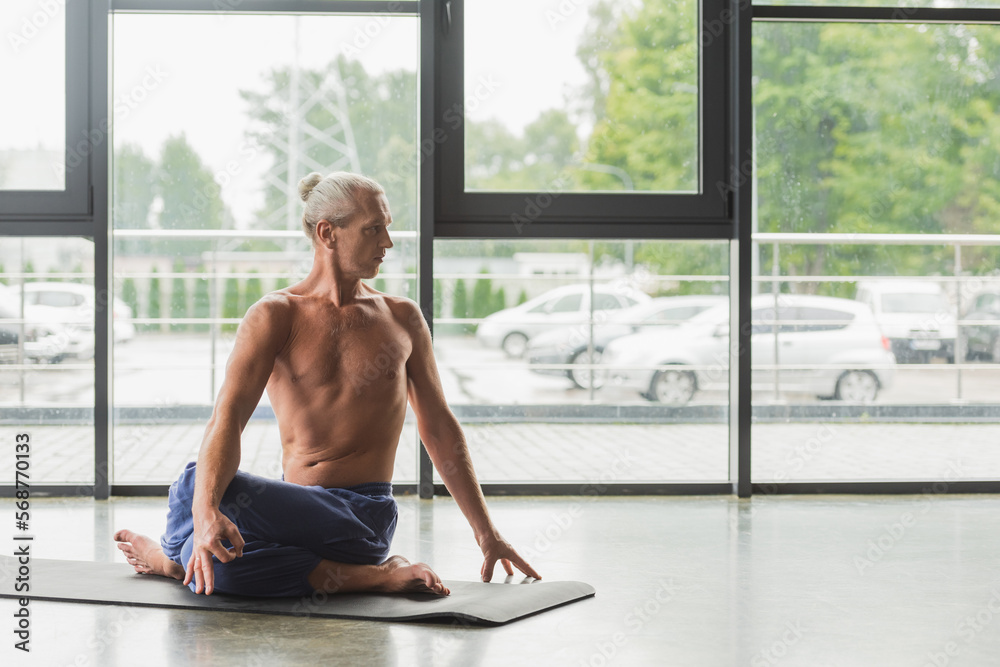 grey haired man in blue pants sitting in twisting yoga pose on mat in studio.
