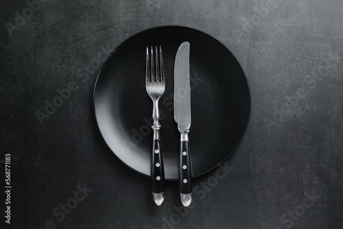Ceramic plate, fork and knife on black table, top view