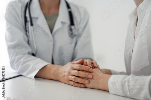 Doctor and patient sitting at the desk in clinic office. The focus is on female physician's hands reassuring woman, close up. Perfect medical service, empathy, and medicine concept.