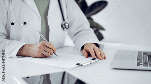 Unknown doctor woman sitting and writing notes at the desk in clinic or hospital office, close up. Medicine concept.