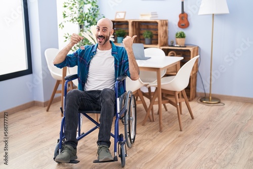 Hispanic man with beard sitting on wheelchair holding new house keys pointing thumb up to the side smiling happy with open mouth
