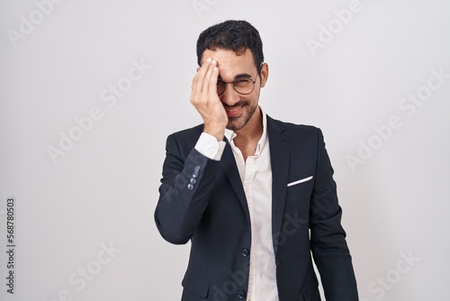 Handsome business hispanic man standing over white background covering one eye with hand, confident smile on face and surprise emotion.