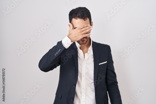 Handsome business hispanic man standing over white background covering eyes with hand, looking serious and sad. sightless, hiding and rejection concept © Krakenimages.com