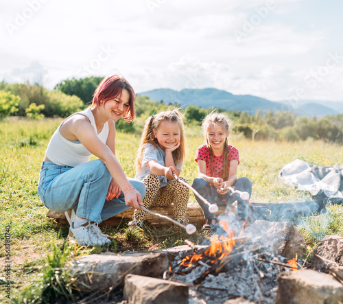 Three sisters cheerfully smiling while they roasting a marshmallows candies on the sticks over the campfire flame. Happy family or outdoor picnic activities concept image..