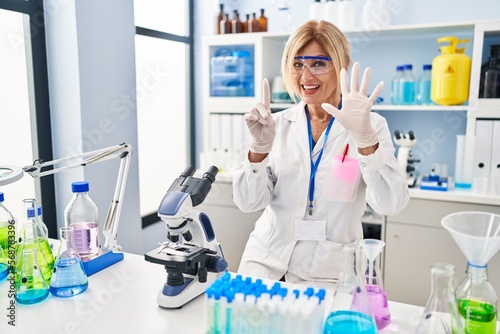 Middle age blonde woman working at scientist laboratory showing and pointing up with fingers number six while smiling confident and happy.
