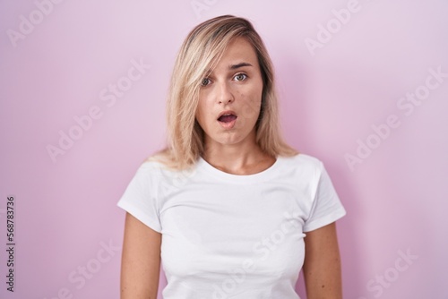 Young blonde woman standing over pink background in shock face, looking skeptical and sarcastic, surprised with open mouth