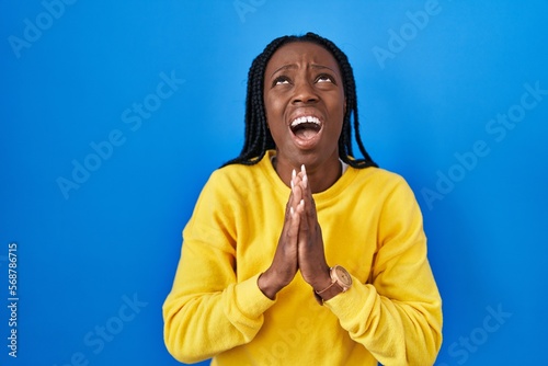 Obraz na plátně Beautiful black woman standing over blue background begging and praying with hands together with hope expression on face very emotional and worried