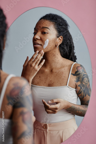 Candid young black woman with acne scars using face cream by mirror