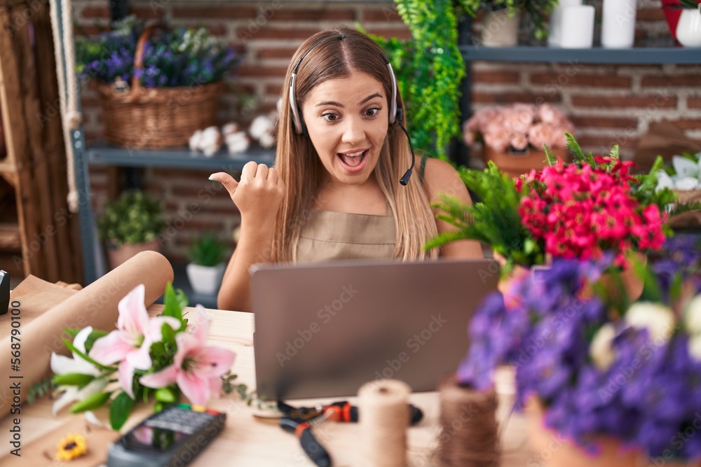 Young blonde woman working at florist shop doing video call pointing thumb up to the side smiling happy with open mouth