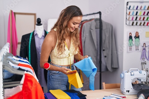 Young beautiful hispanic woman tailor smiling confident holding cloths at tailor shop