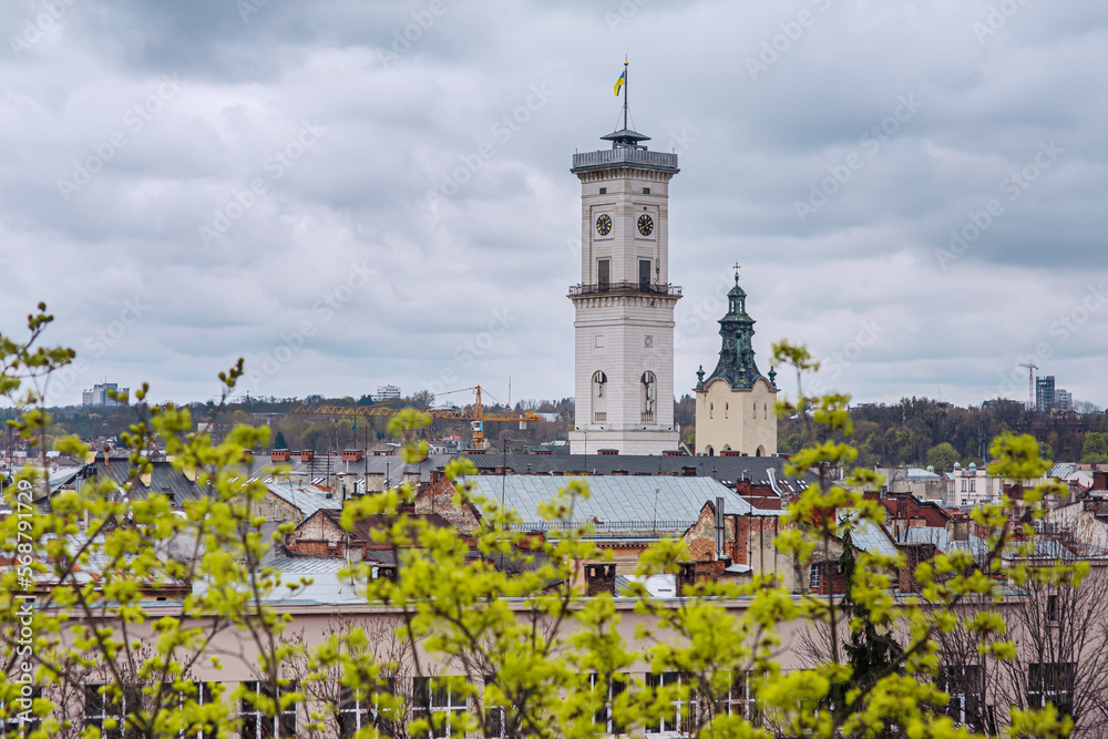Scenic view of centre of Lviv (Ukraine) old town, city hall tower with flag and clock, roofs, church spire - famous tourist landmarks on overcast rainy day with clouds, spring trees in the foreground