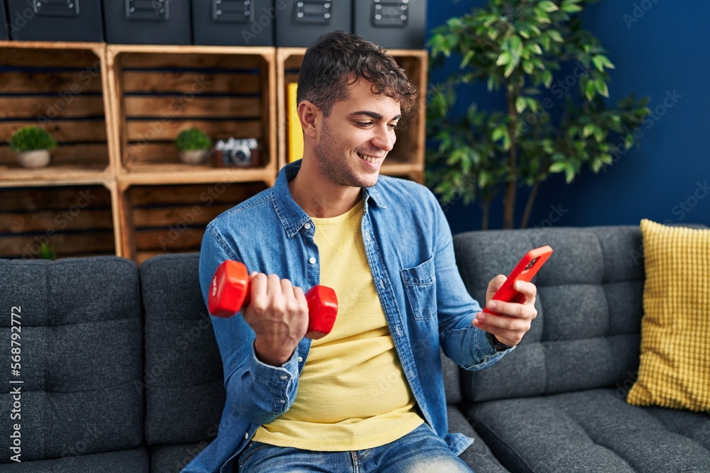 Young hispanic man using smartphone training with dumbbell at home Young hispanic man using smartphone training with dumbbell at home