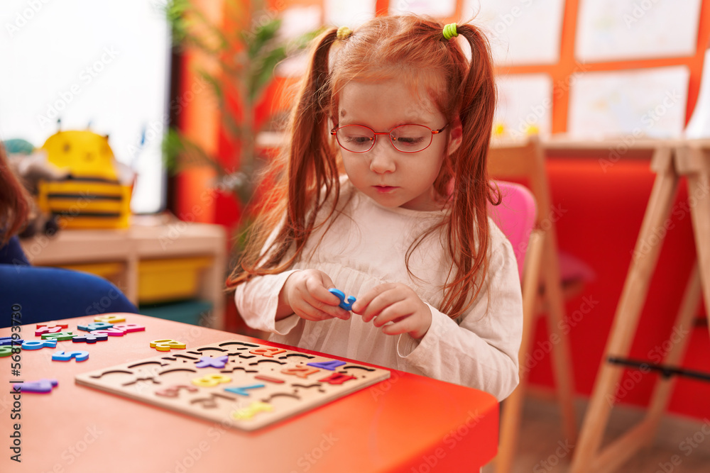 Fototapeta premium Adorable redhead girl playing with maths puzzle game sitting on table at kindergarten
