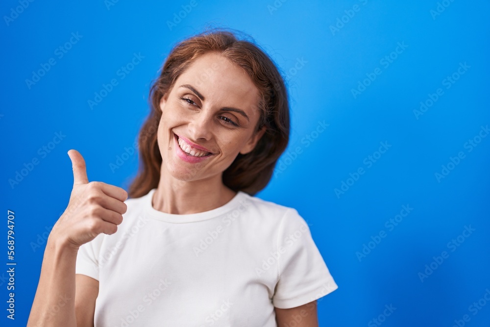 Young beautiful hispanic woman smiling confident doing ok sign with thumb up over isolated blue background Young beautiful hispanic woman smiling confident doing ok sign with thumb up over isolated blue background