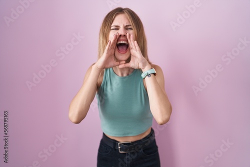 Blonde caucasian woman standing over pink background shouting angry out loud with hands over mouth