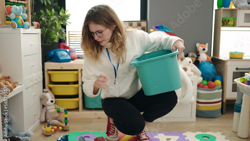 Young blonde woman preschool teacher picking up toys at kindergarten photo