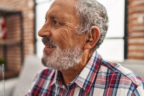 Senior grey-haired man using hearing aid sitting on sofa at home