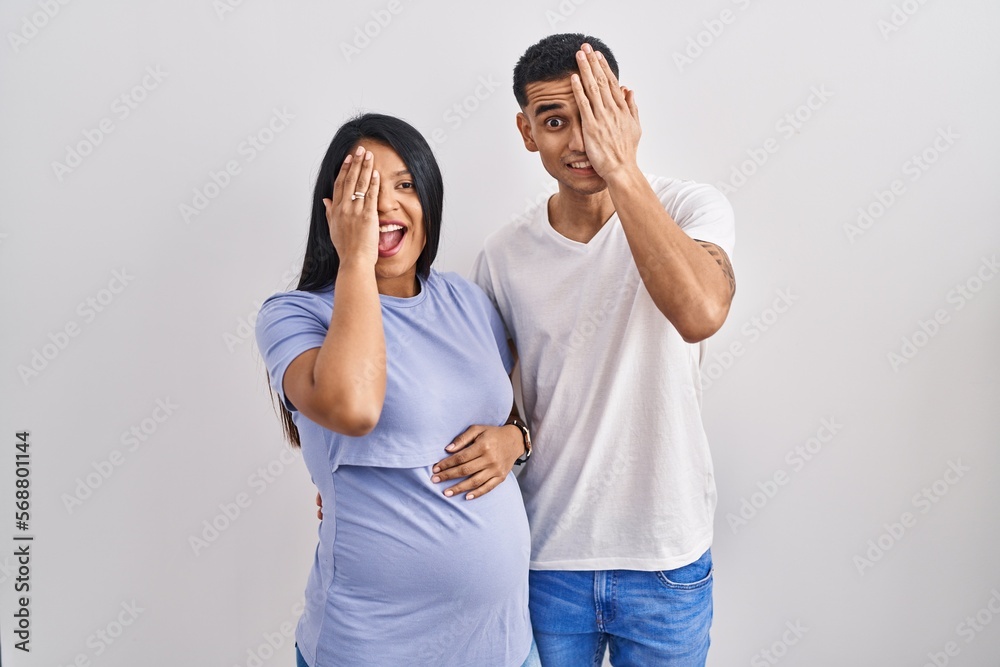 Young hispanic couple expecting a baby standing over background covering one eye with hand, confident smile on face and surprise emotion. Young hispanic couple expecting a baby standing over background covering one eye with hand, confident smile on face and surprise emotion.