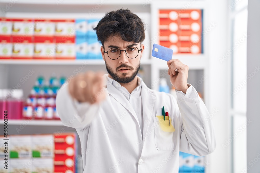 Hispanic man with beard working at pharmacy drugstore holding credit card pointing with finger to the camera and to you, confident gesture looking serious Hispanic man with beard working at pharmacy drugstore holding credit card pointing with finger to the camera and to you, confident gesture looking serious