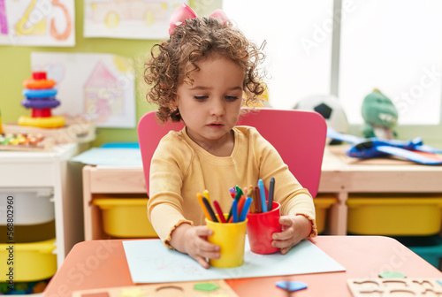 Adorable hispanic toddler student sitting on table drawing on paper at kindergarten